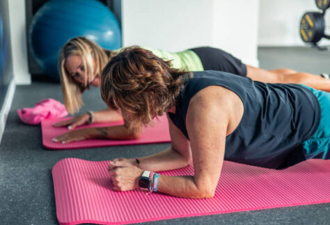 Two older women, dressed in sportswear, are performing planks on their yoga mats in a gym. The image highlights their dedication to fitness and strength training, focusing on core muscles